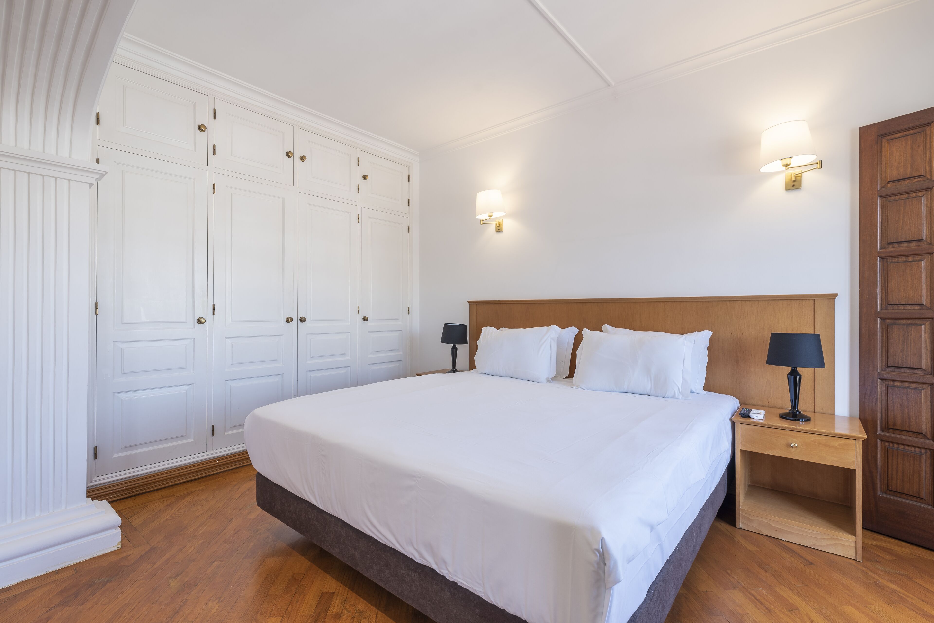 Una cama bien hecha con sábanas blancas, una mesita de noche de madera con una lámpara y un amplio armario con múltiples puertas.