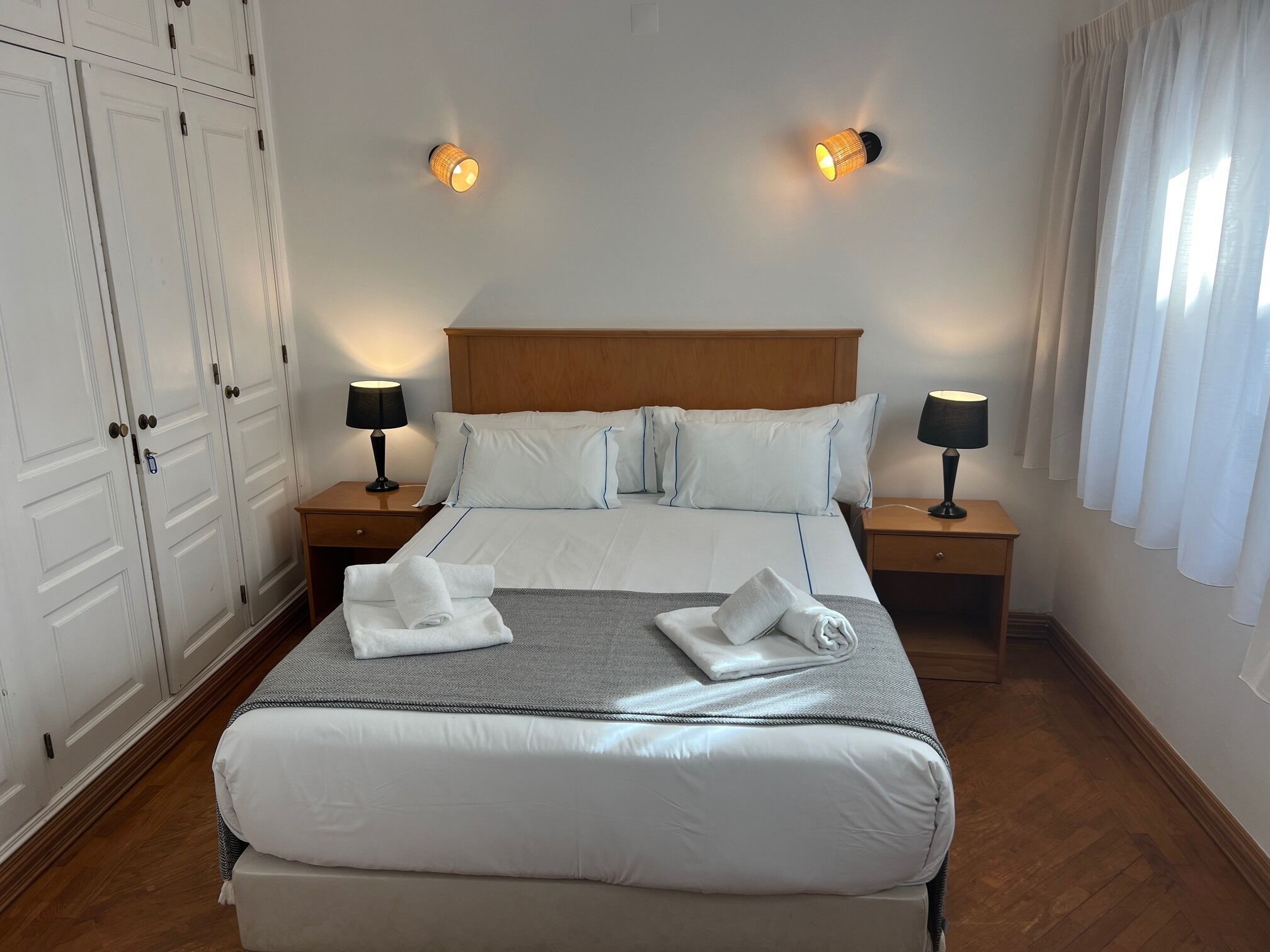 A hotel room with a large bed, two bedside tables with lamps, a wooden headboard, white curtains, and a wardrobe with multiple doors.