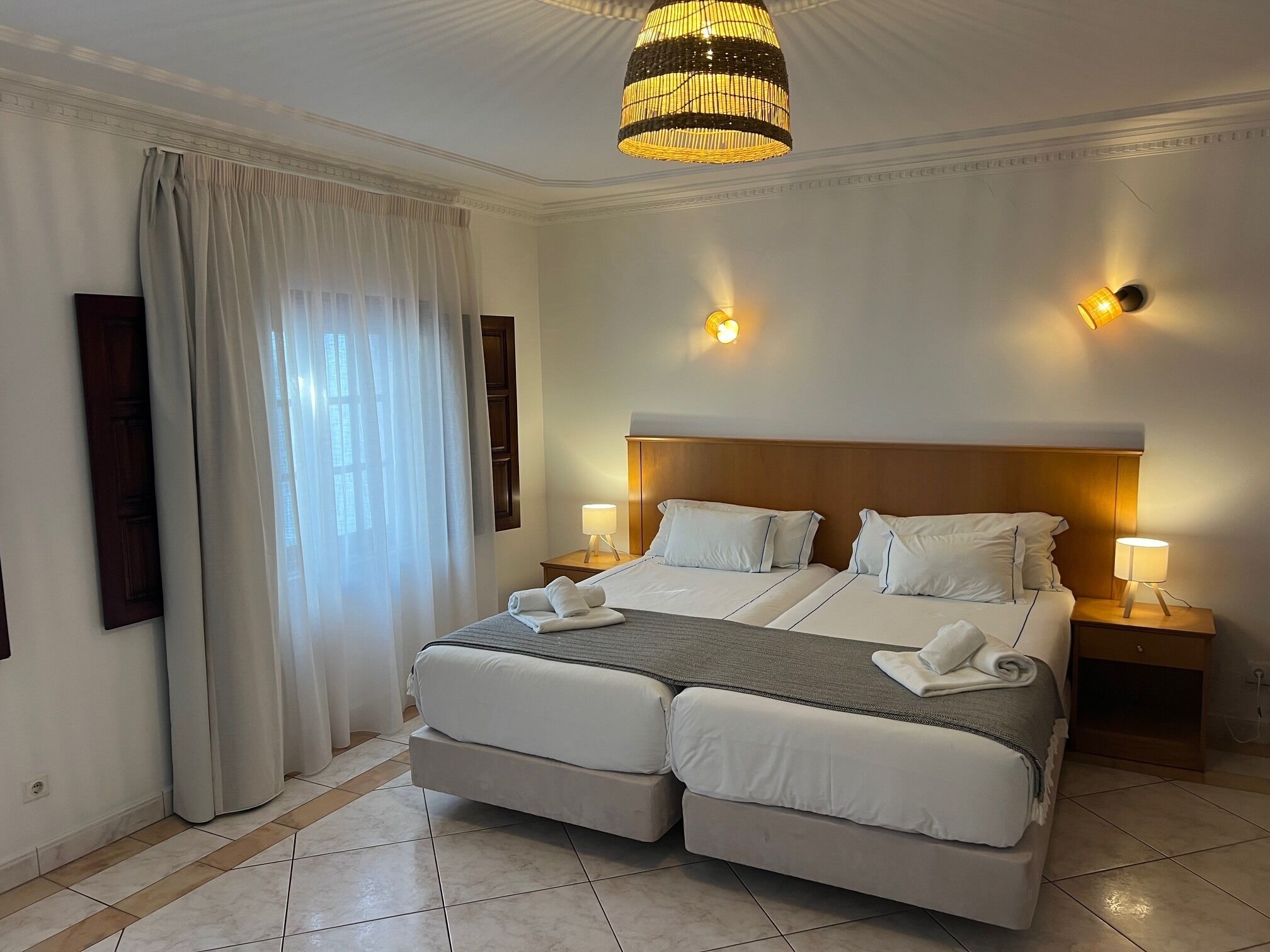 A double bed with two nightstands, a chandelier, and a large window with curtains.