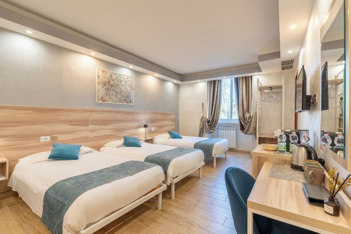 A hotel room with three beds, a desk with a chair, a mirror, and a window with curtains.