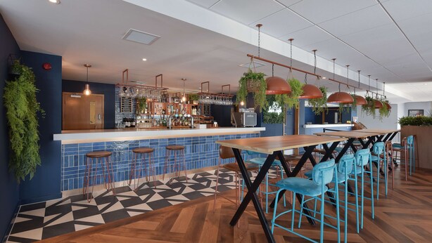 Een moderne bar met een houten toonbank, blauwe betegelde achterwand en hangende planten. Er staan barstoelen en een lange houten tafel met blauwe stoelen.