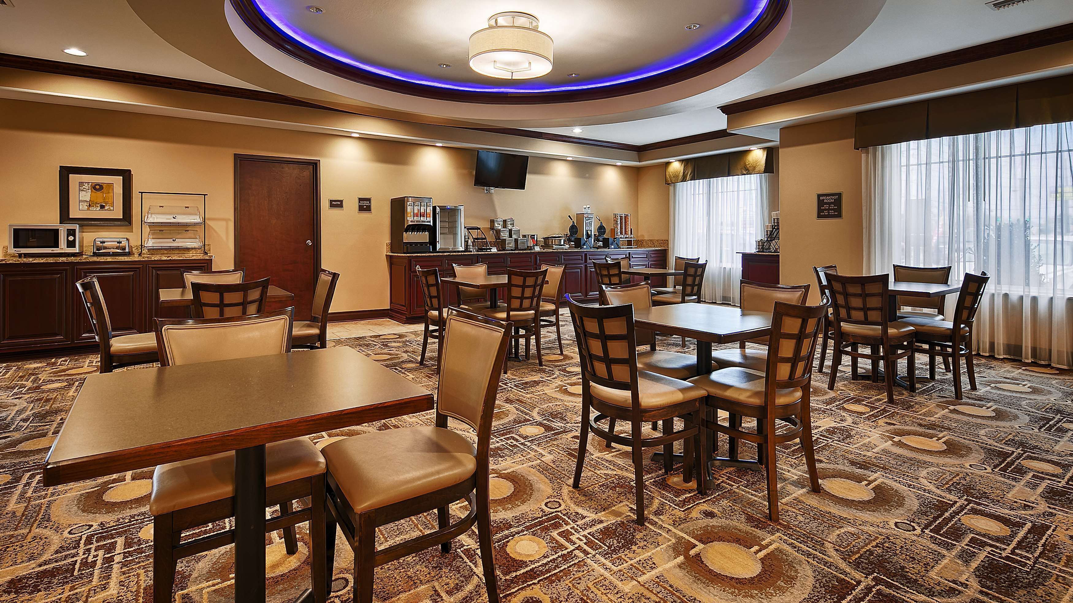 A hotel dining area with multiple tables and chairs, a kitchen area with a coffee station, and a television mounted on the wall.