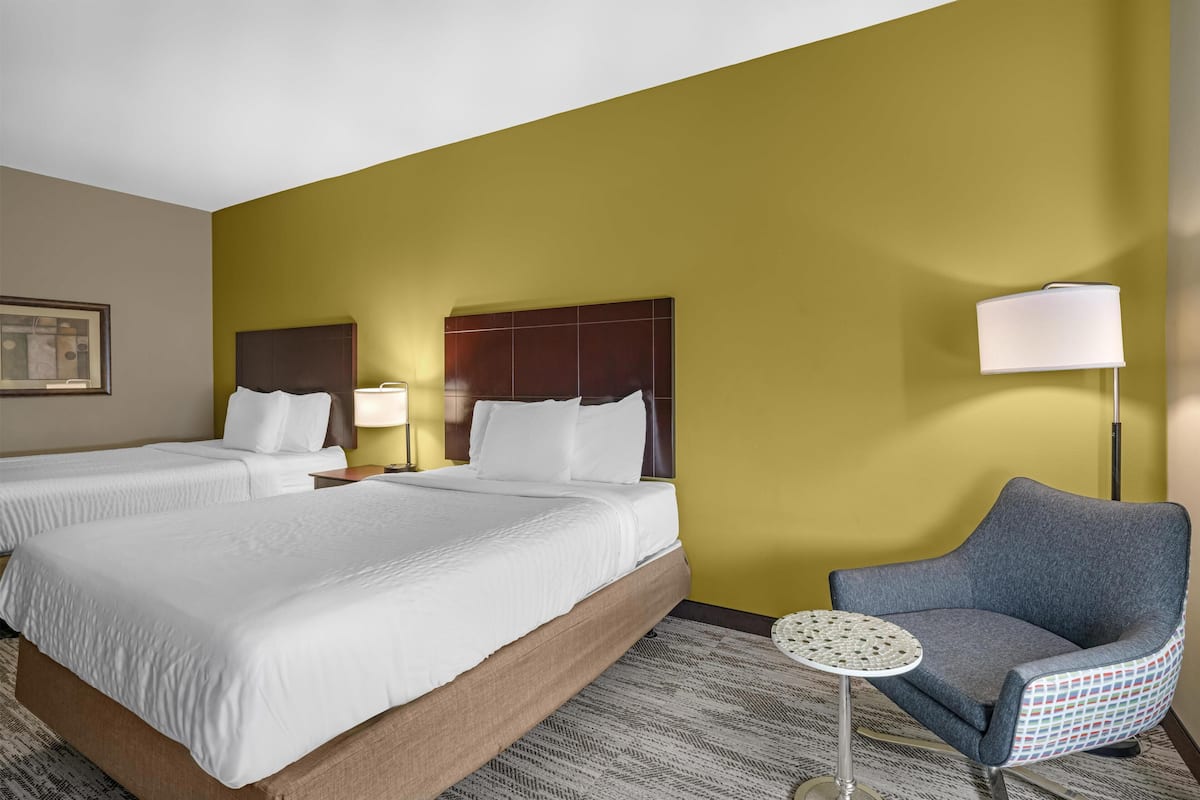 A hotel room with two beds, a small round table, a chair, and a lamp.