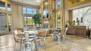 A spacious indoor area with stone flooring, a stone fireplace, a television mounted on the wall, and a bar area with stools and a refrigerator. - Raintree at Phoenix South Mountain Preserve (Phoenix)