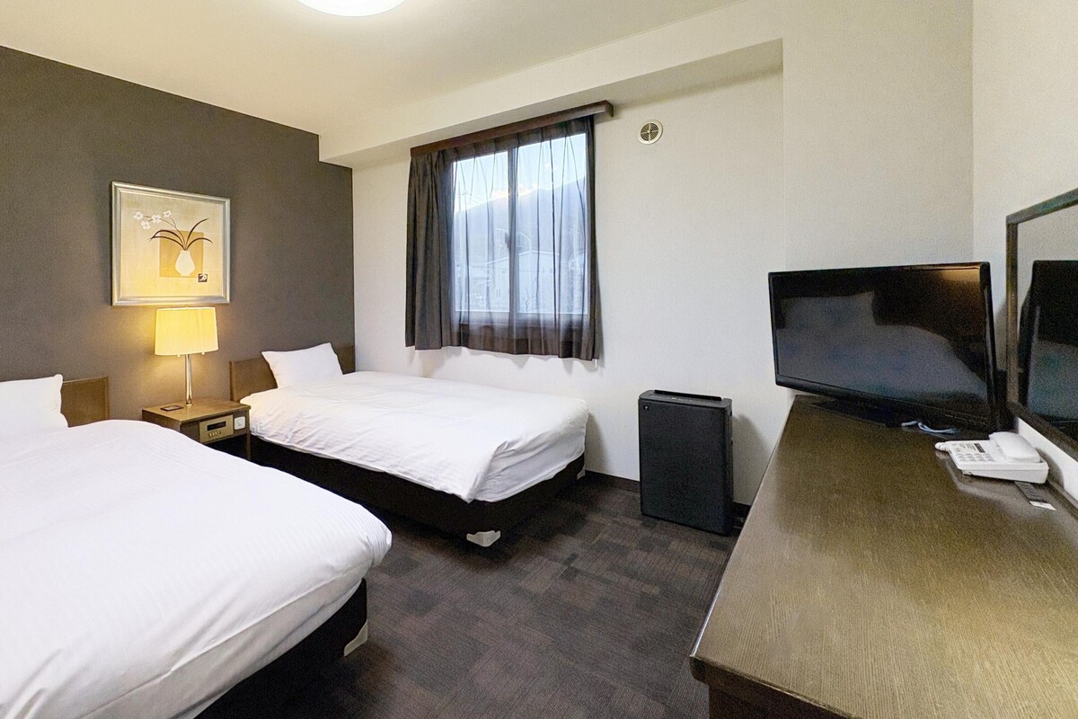 Habitación de hotel con dos camas, un escritorio, televisión, una lámpara y una ventana con cortinas.