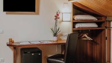 A compact room with a wooden ceiling, a desk with a computer and papers, a chair, a small fridge, and a closet with hanging clothes.