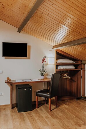 A compact room with a wooden ceiling, a desk with a computer and papers, a chair, a small fridge, and a closet with hanging clothes. - Hotel Arrope (Haro)