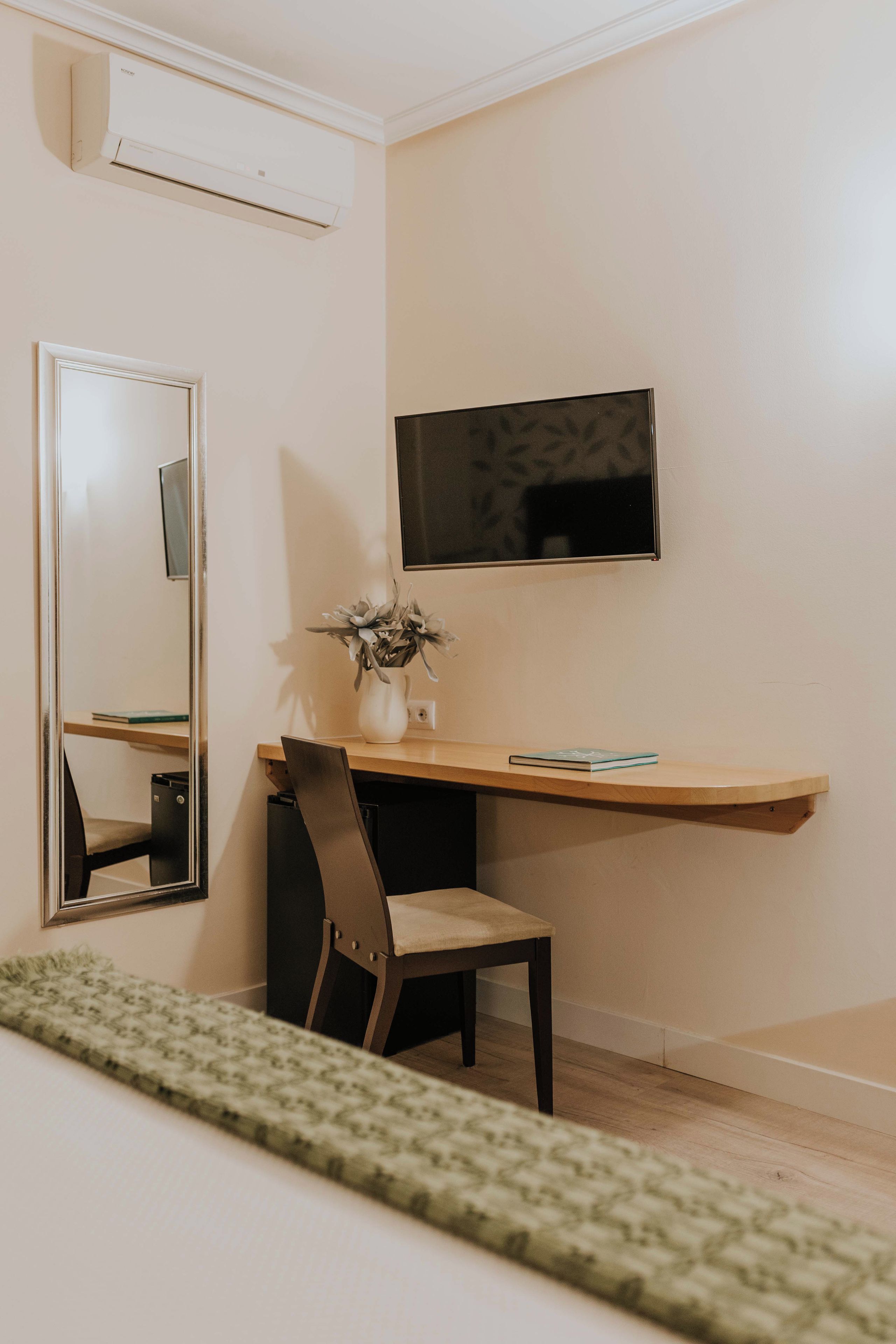 A room with a desk, chair, mirror, and a flat-screen TV mounted on the wall.
