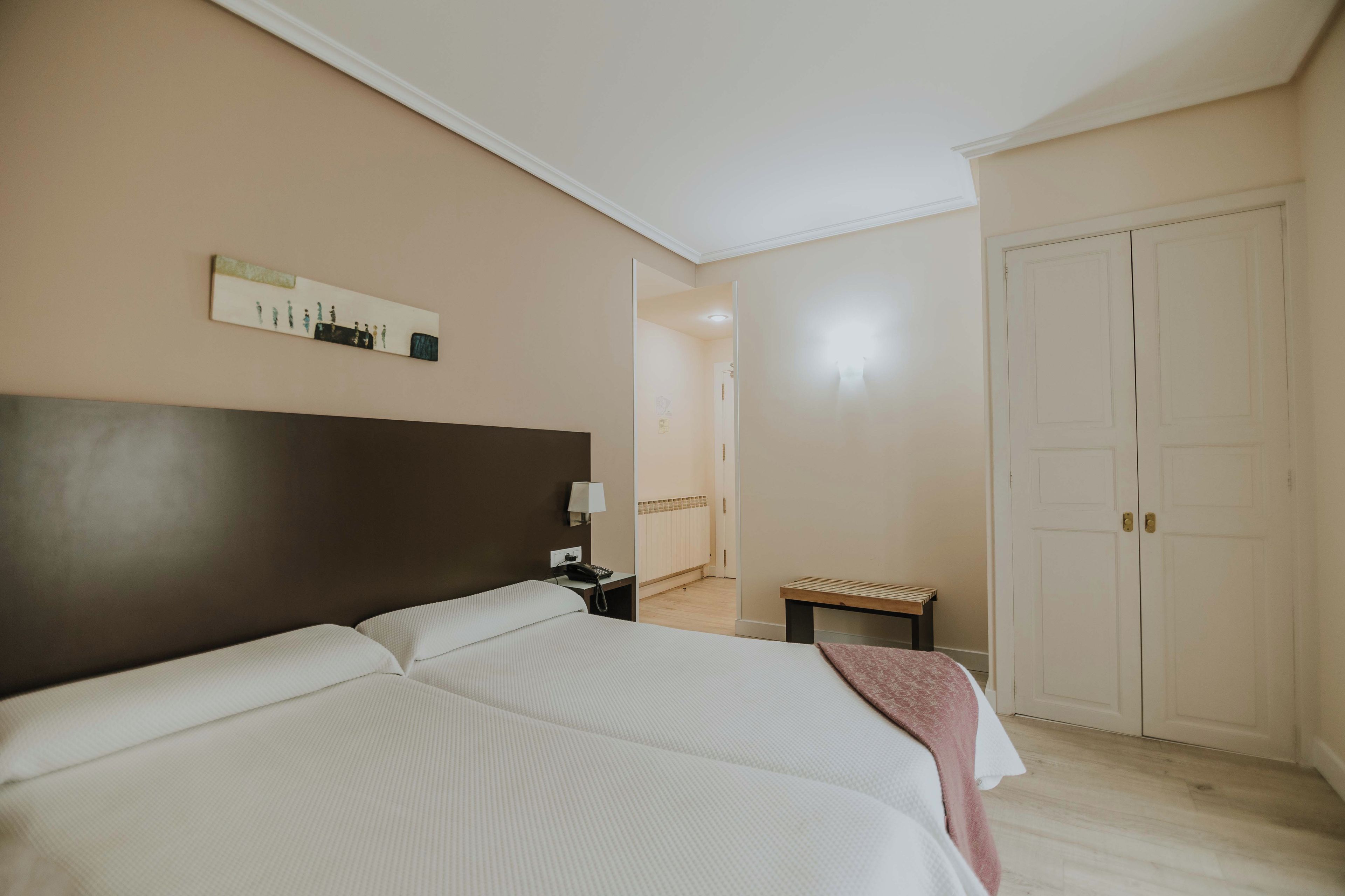 Een hotelkamer met een groot bed, een nachtkastje met een lamp, een klein bankje en een deur.