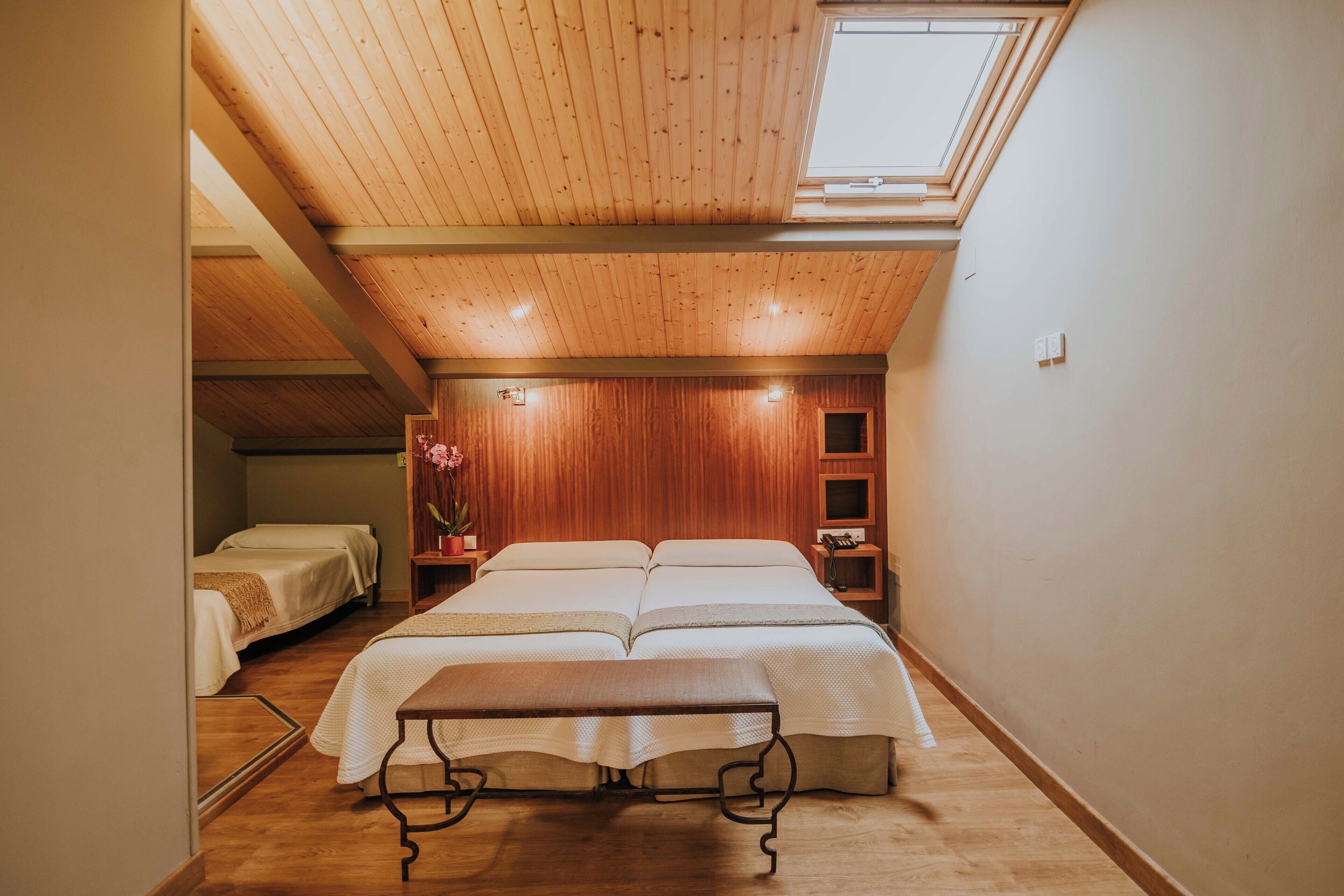 A bedroom with a wooden ceiling, a skylight, a large bed, bedside tables, and a small bedside lamp.
