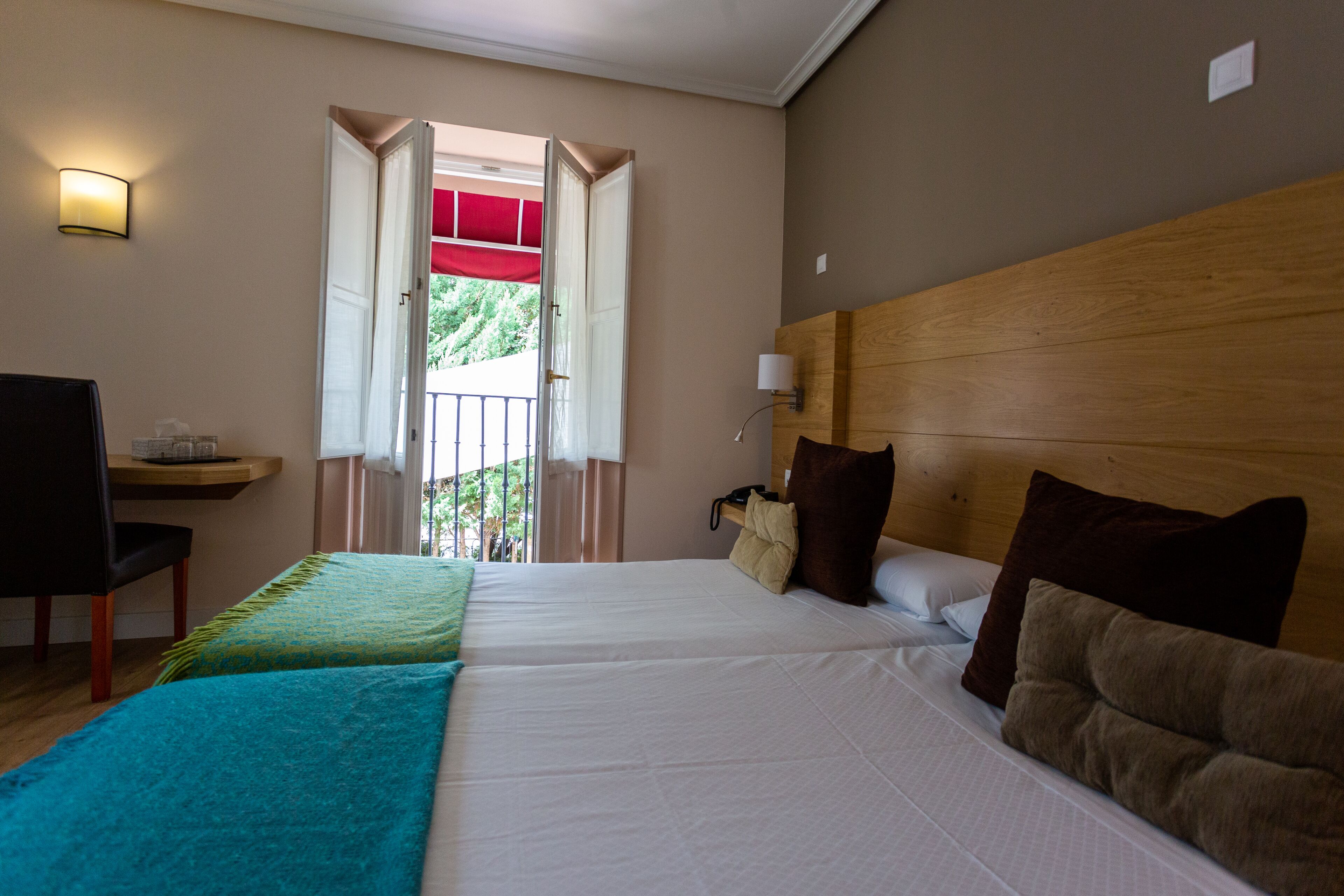 Quarto de hotel com duas camas, uma escrivaninha com cadeira, mesa de cabeceira e janela com porta aberta.