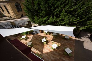 An outdoor patio area with tables and chairs, surrounded by a building and a large tree.