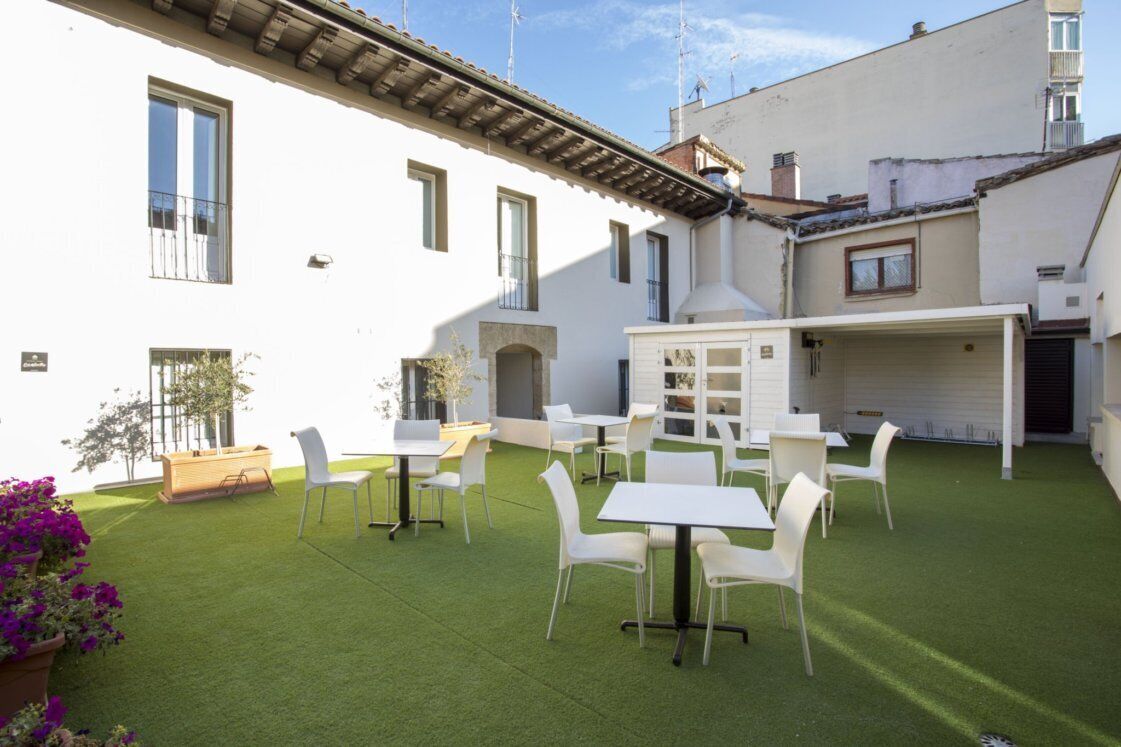 Un patio con césped artificial, mesas y sillas blancas, y una pequeña estructura de madera.