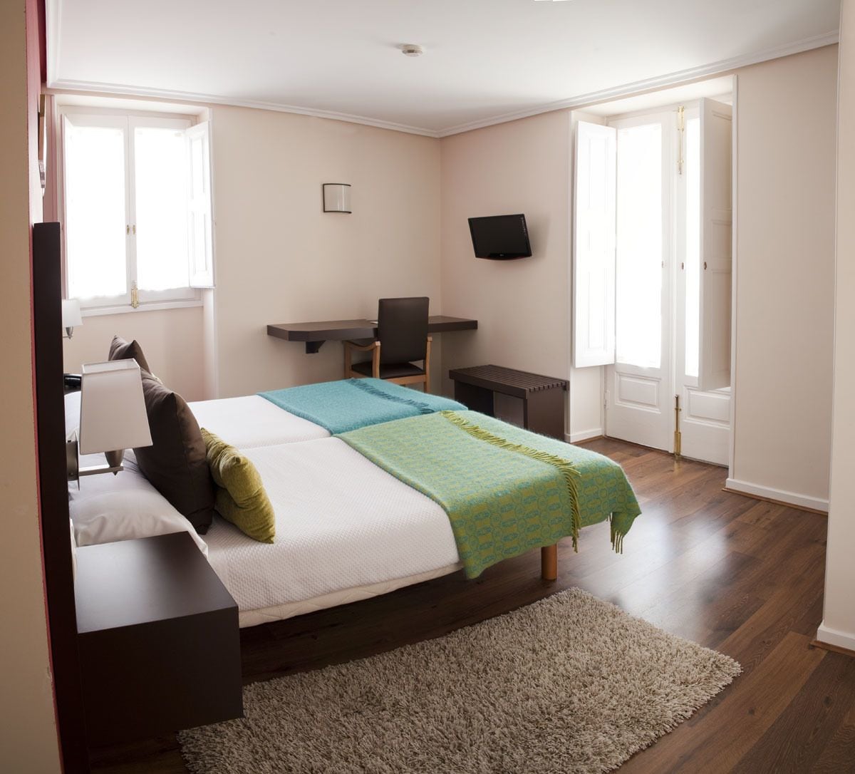 A hotel room with a bed, desk, chair, TV, and large windows.