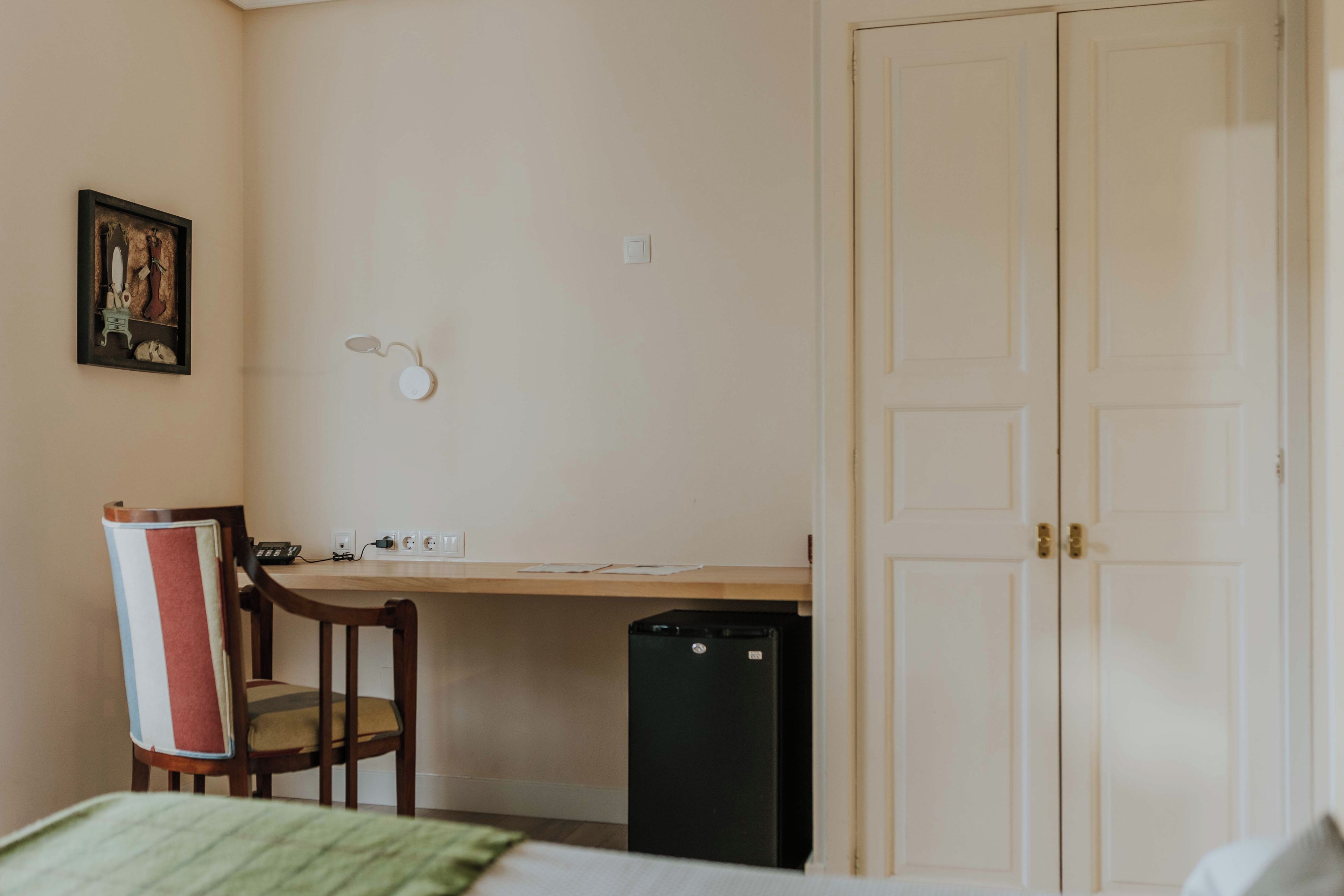 Una habitación con cama, escritorio, silla, un cesto de basura pequeño y una puerta.