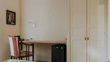 Una habitación con cama, escritorio, silla, un cesto de basura pequeño y una puerta.