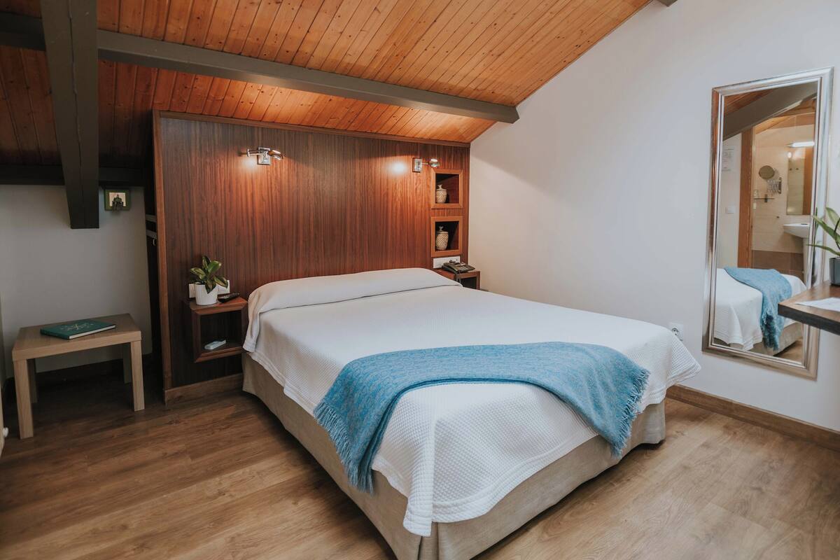 A bedroom with a wooden ceiling, a bed with a white and blue blanket, a bedside table with a book, and a mirror on the wall.