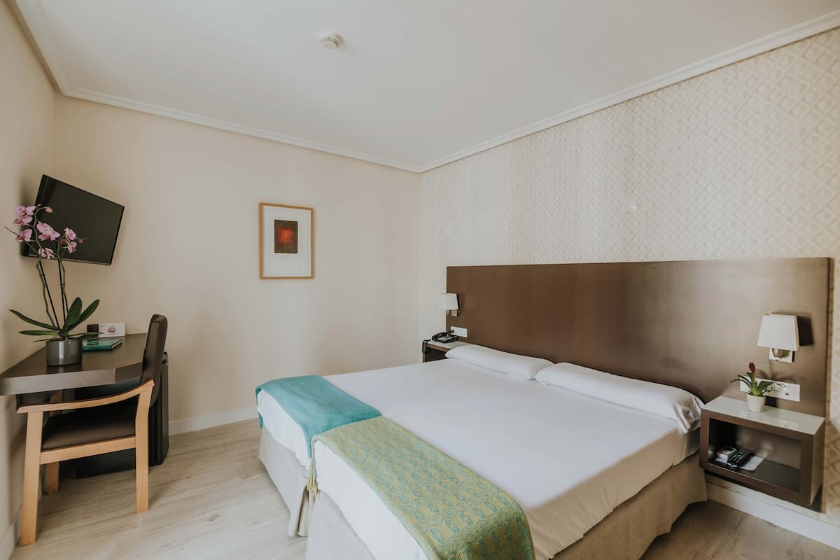 A hotel room with a bed, bedside table, lamp, and a desk with a chair and a potted plant.