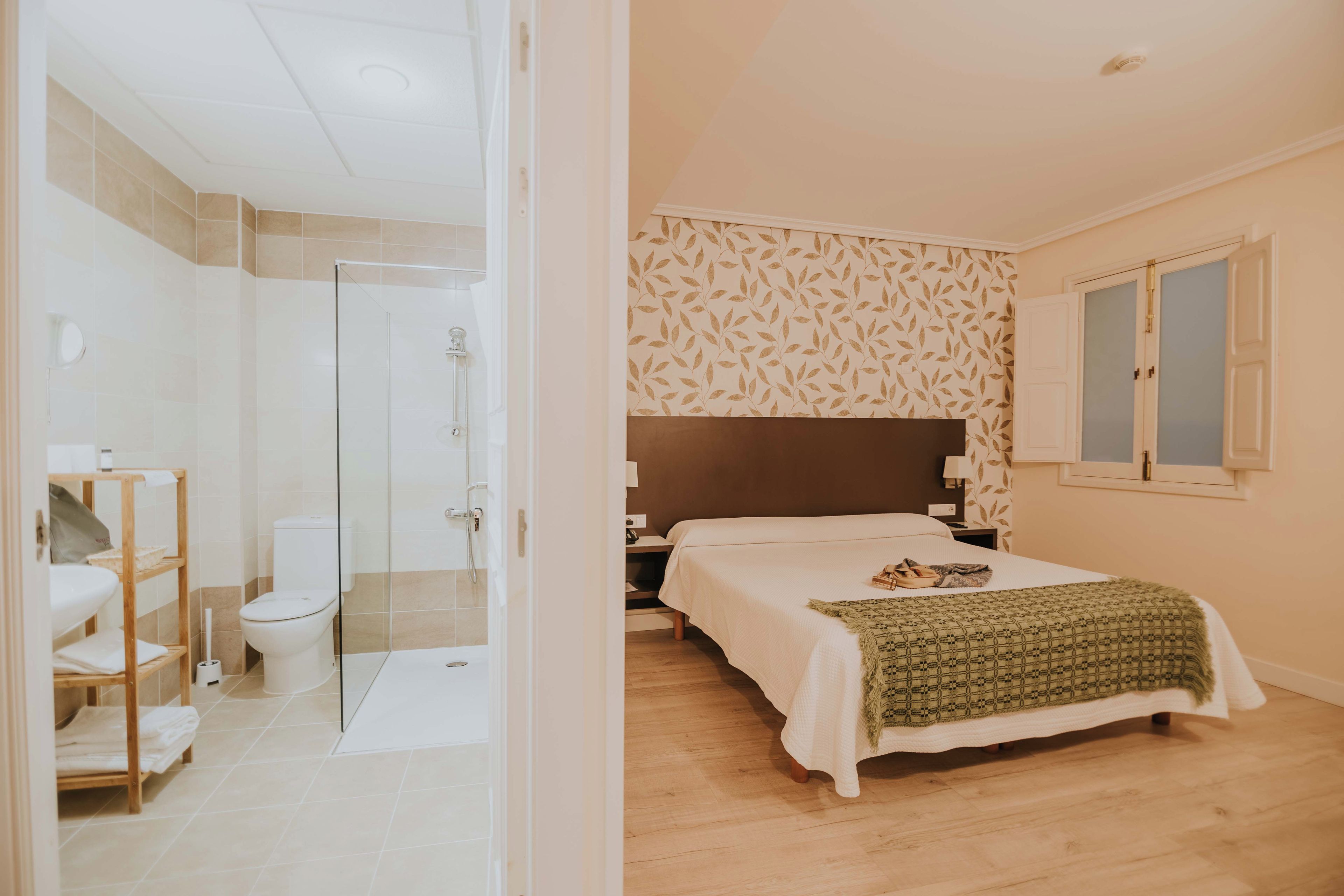 A bedroom with a bed, a nightstand, a window, and a bathroom with a shower and toilet.