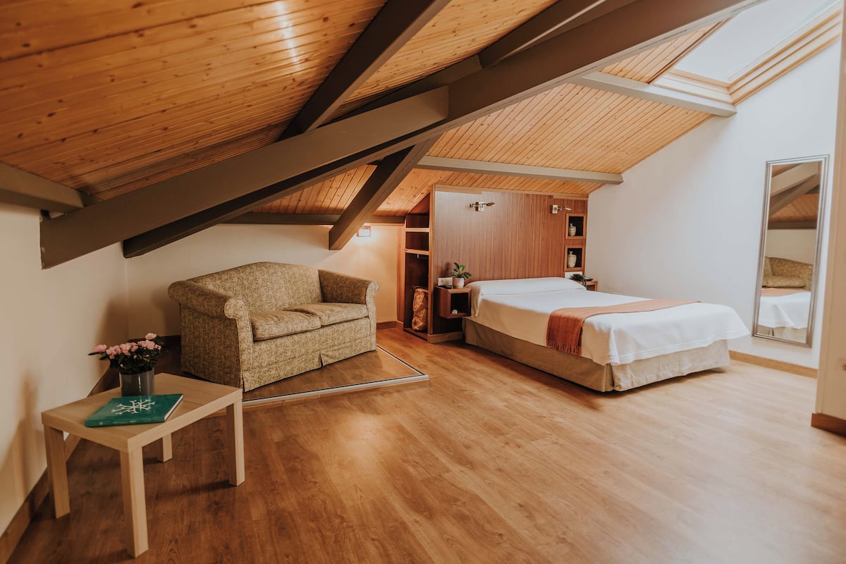 A bedroom with a wooden ceiling, a bed, a sofa, and a small table with a book and flowers.