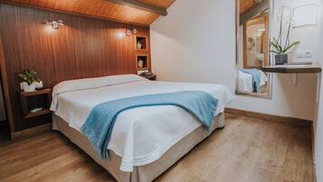 A bedroom with a wooden ceiling, a bed with a white and blue blanket, a bedside table with a plant, and a mirror on the wall.