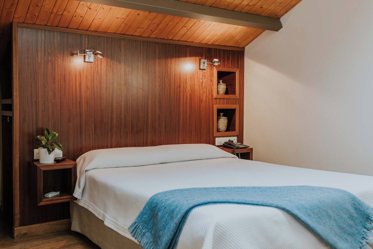 A bedroom with a wooden headboard, a bed with white bedding, a blue throw blanket, and two bedside tables with lamps and a plant.