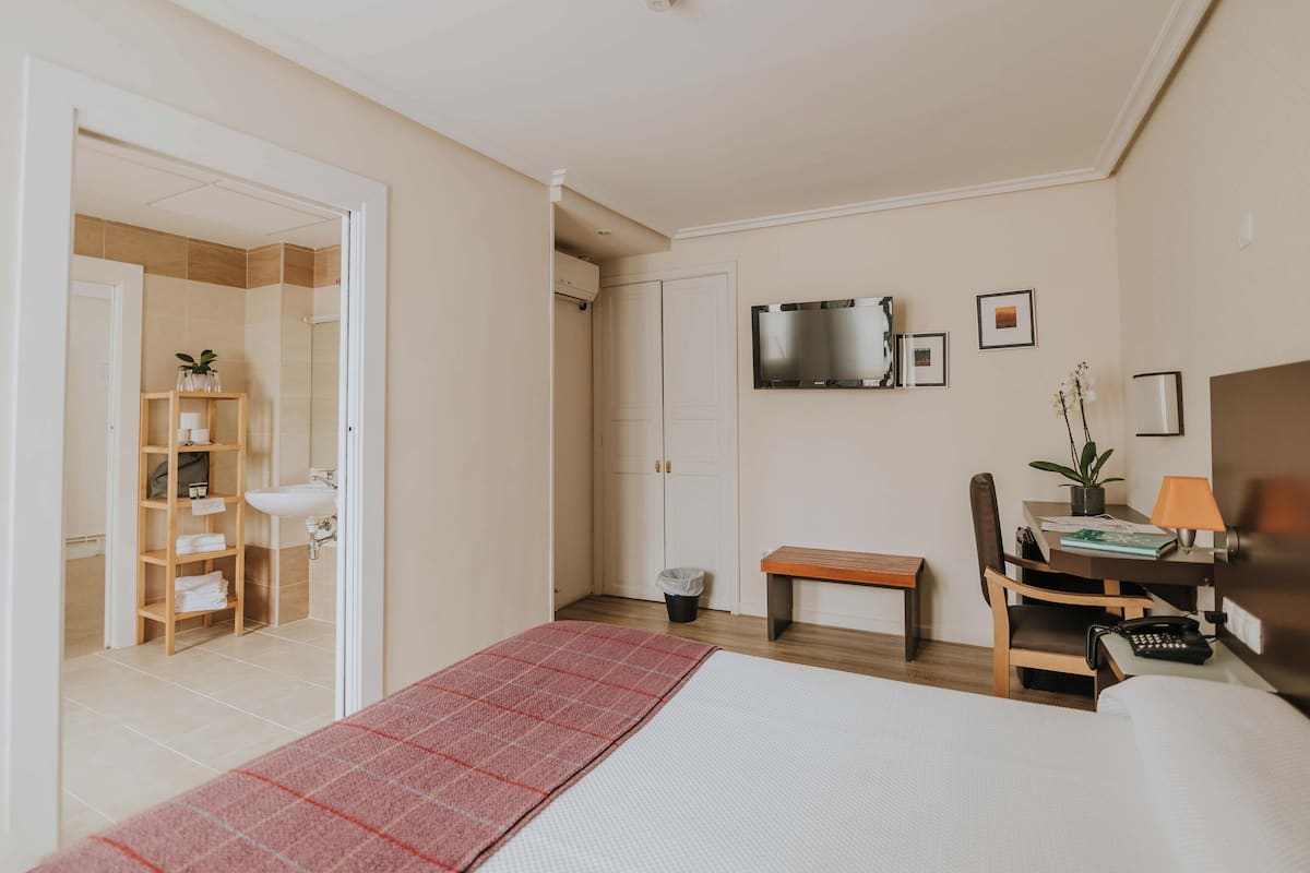 A hotel room with a bed, a desk with a chair, a TV, and a bathroom visible through an open door.