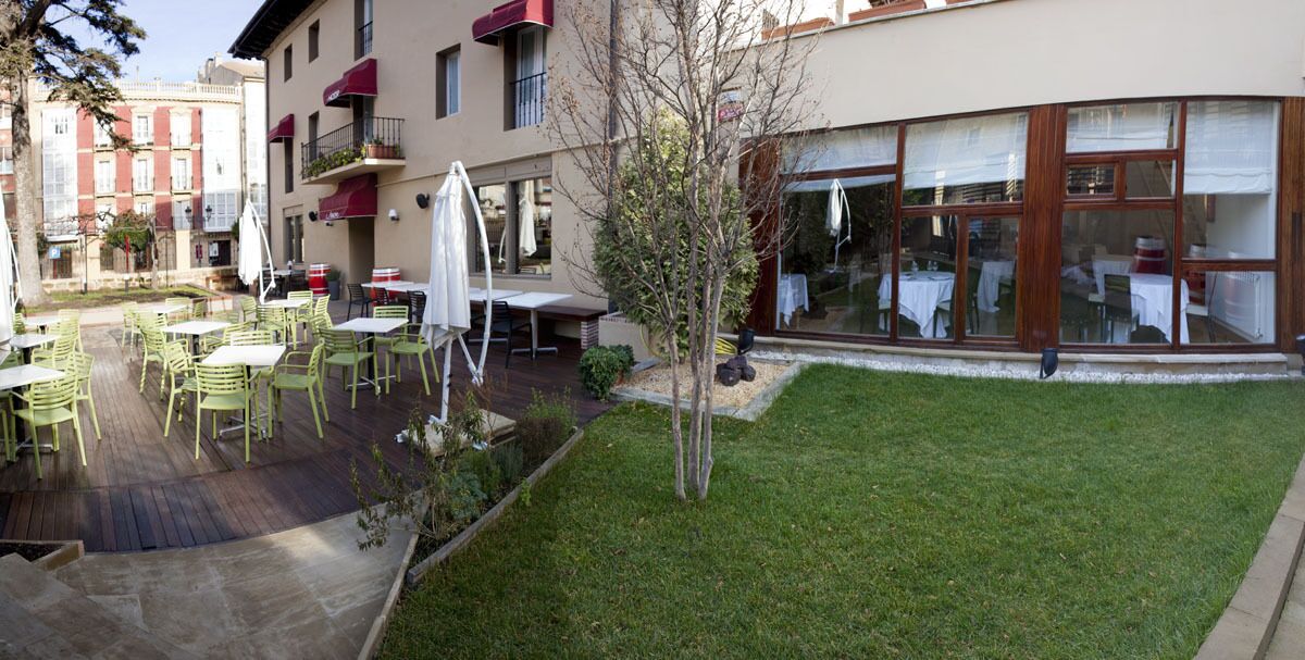 Eine Terrasse im Freien mit Tischen und Stühlen, ein kleiner Baum und ein Gebäude mit großen Fenstern und einer roten Markise.