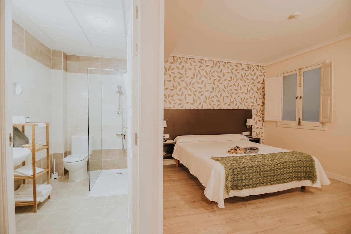 A bedroom with a bed, a nightstand, a window, and a bathroom with a shower and toilet.