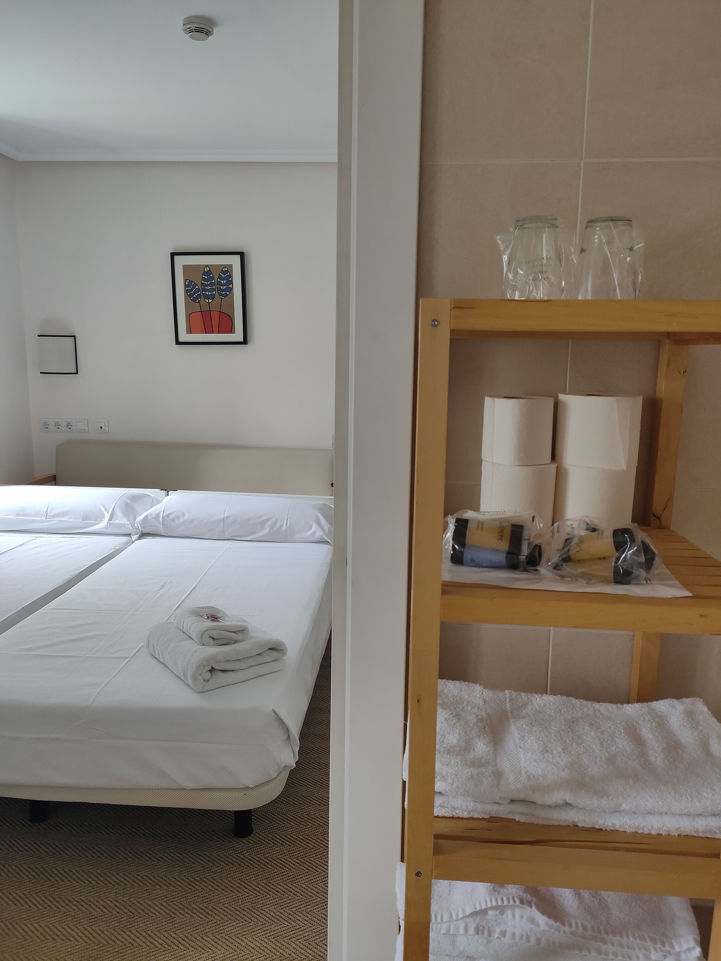 Ein Hotelzimmer mit zwei Betten, einem Holzboden mit Handtüchern und Toilettenartikeln, einem gerahmten Bild an der Wand und einer Deckenleuchte.