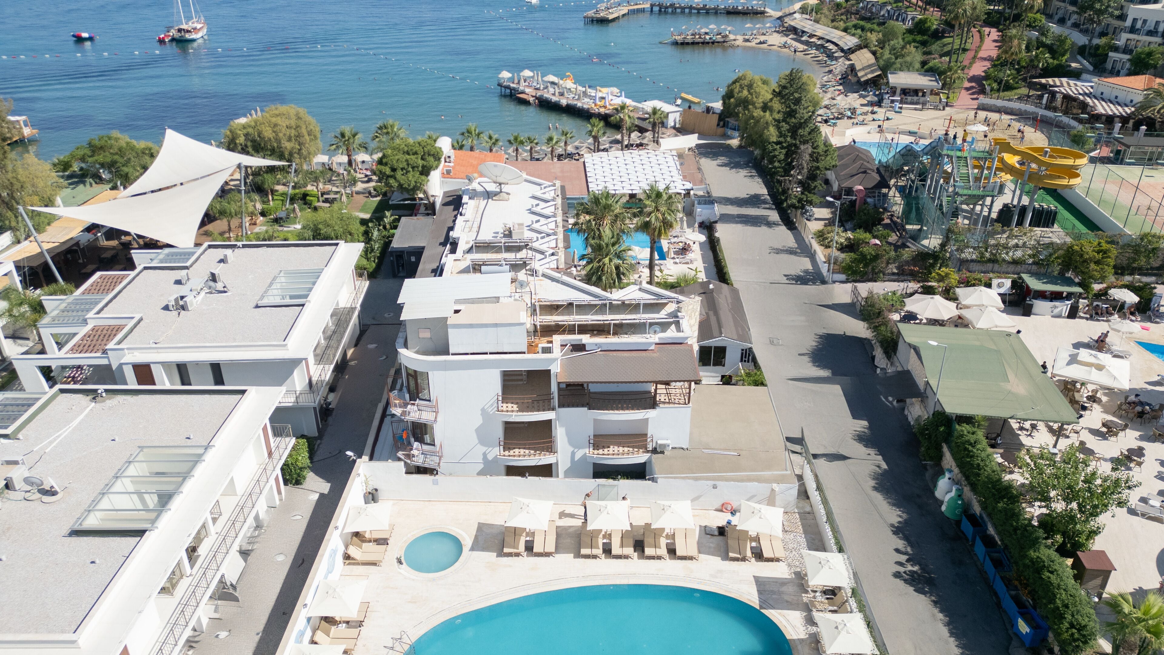 A modern hotel with a rooftop pool, a marina with boats, and a beach area with umbrellas and sun loungers.