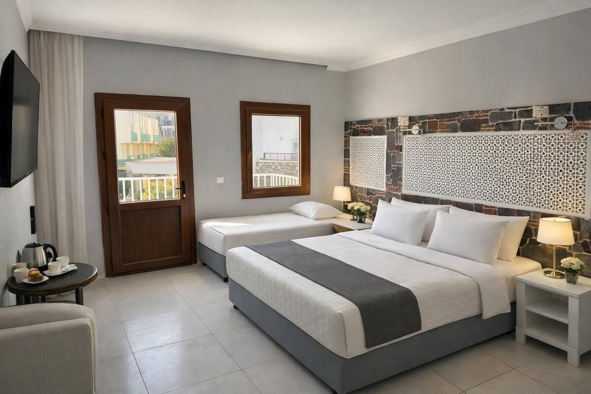 Ein Hotelzimmer mit zwei Betten, einem Fernseher, einem kleinen Tisch mit Wasserkocher und Blick auf einen Balkon.