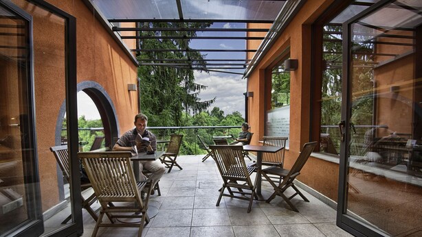 An outdoor patio with wooden chairs and tables, large windows, and a view of greenery. - Cukrarna Hostel (Kranj)
