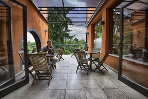 An outdoor patio with wooden chairs and tables, large windows, and a view of greenery. - Cukrarna Hostel (Kranj)