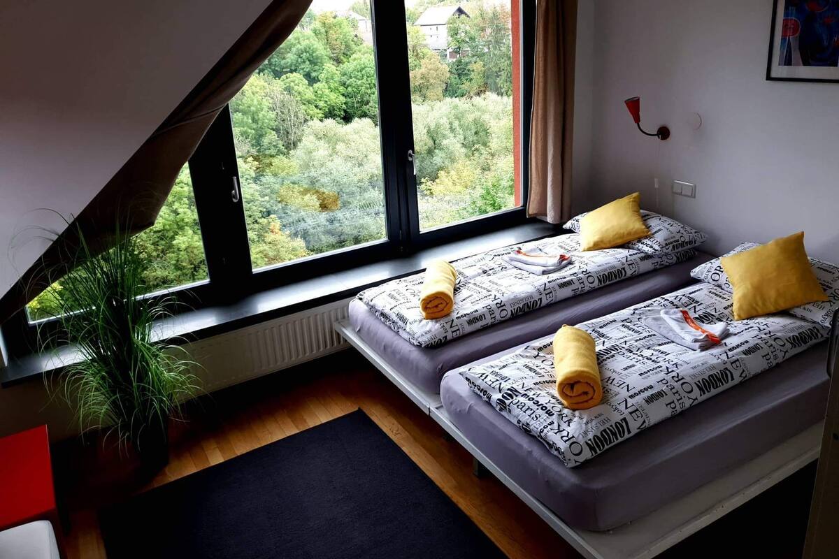 Quarto com teto inclinado, duas camas com roupa de cama estampada e almofadas amarelas, um vaso com planta e vista para o verde lá fora.