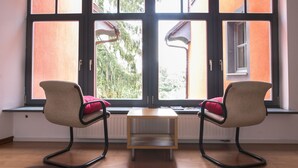 A room with two chairs, a small table, and a large window with a view of trees and a building outside. - Cukrarna Hostel (Kranj)