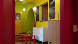 A brightly lit room with a yellow wall, a white sofa, a red table, and a chalkboard menu. - Cukrarna Hostel (Kranj)