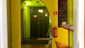 A hallway with a green archway leading to a door marked "IZHOOD". A chalkboard menu lists coffee, beer, wine, ice tea, coca cola, and coca cola. - Cukrarna Hostel (Kranj)