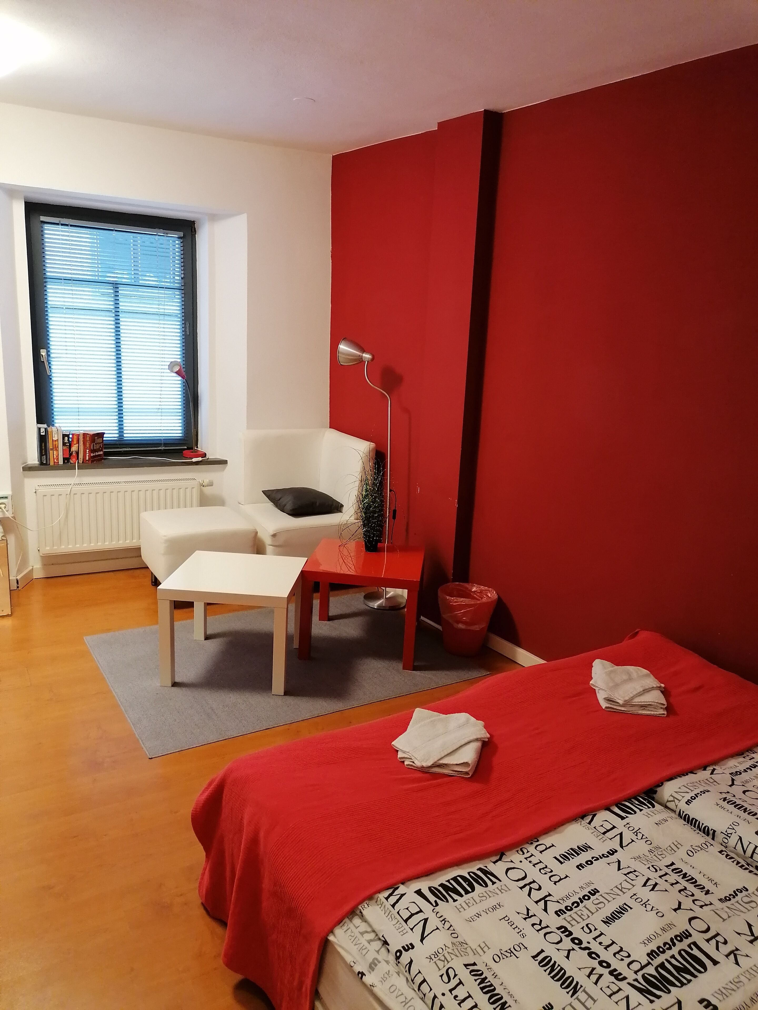 A bedroom with a bed, a desk, a chair, a radiator, a window with blinds, and a red wall.