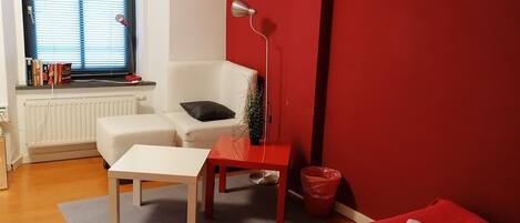 A bedroom with a bed, a desk, a chair, a radiator, a window with blinds, and a red wall.