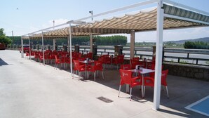 An outdoor café with red chairs and tables, a covered seating area, and a view of a river and hills. - Hotel Alameda (Alba de Tormes)