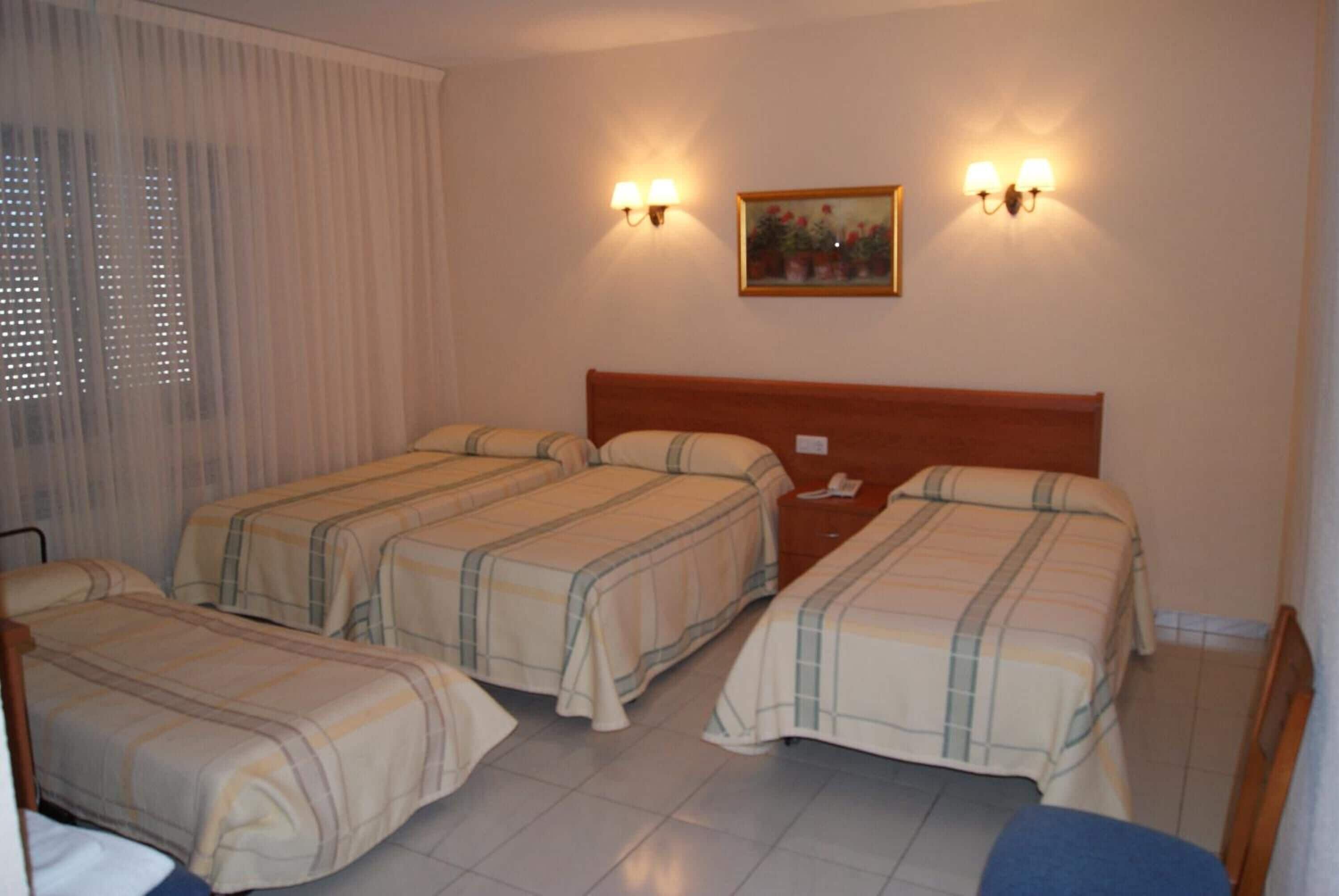 Habitación de hotel con tres camas, cabecera de madera, dos lámparas fijadas a la pared y un cuadro.