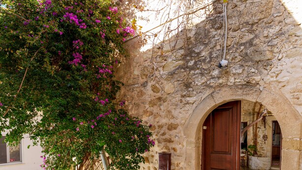 Un edificio de piedra con una puerta de madera y un pequeño arco. Un árbol con flores moradas está frente al edificio.