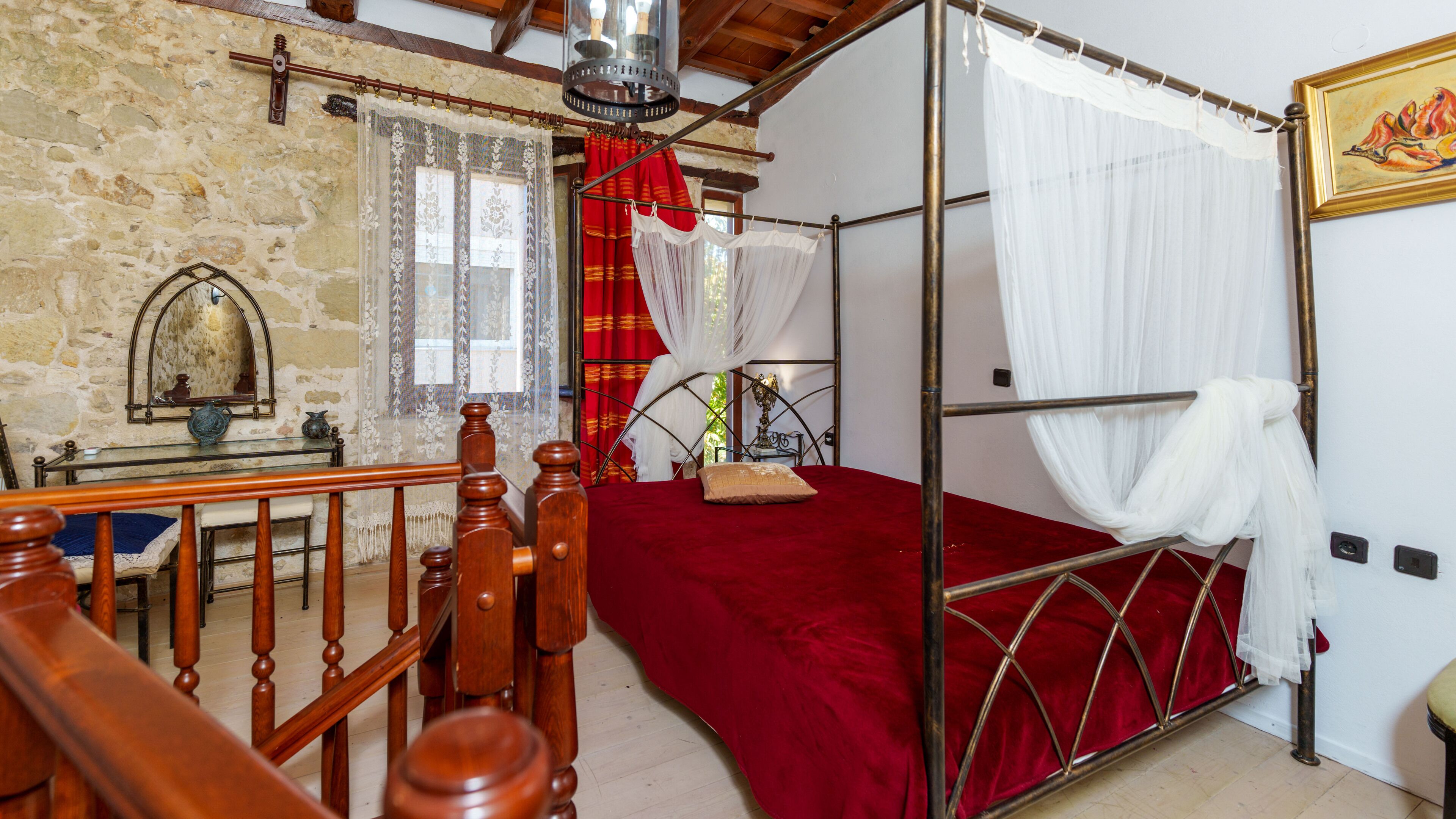 Un dormitorio con pared de piedra, estructura de cama de madera, colcha roja, cama con dosel, silla de madera, mesa con un florero y un cuadro en la pared.