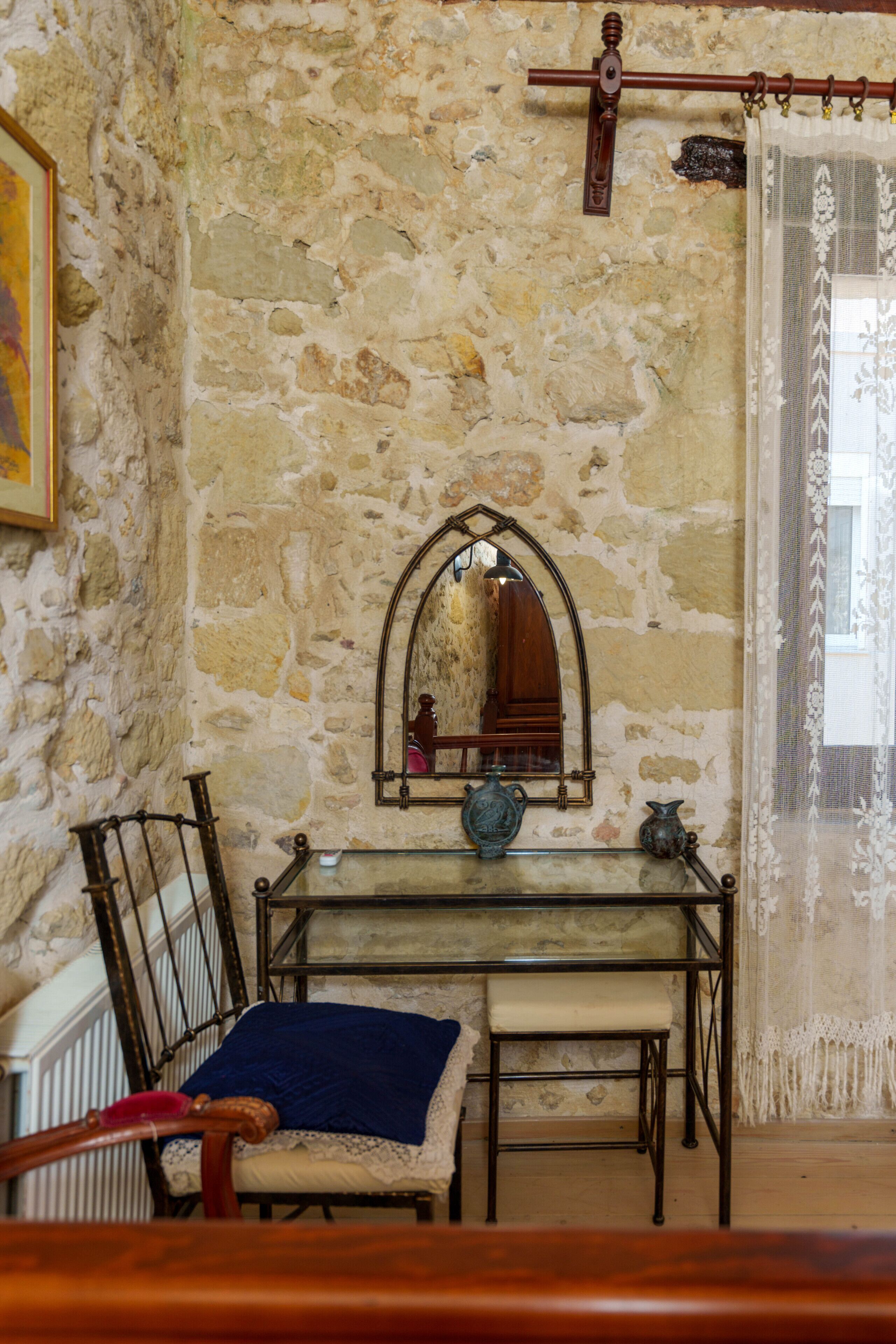 Quarto com paredes de pedra, um banco de madeira, uma mesa pequena com um vaso e um espelho na parede.