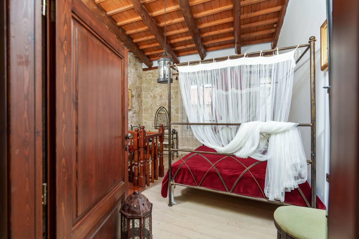Ein Schlafzimmer mit Holzbalkendecke, Metallbettgestell, roter Bettdecke und weißem Baldachin.
