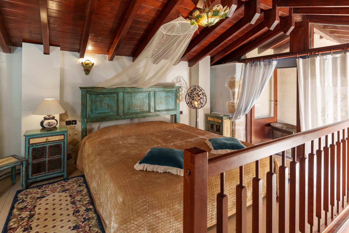 Ein Schlafzimmer mit einem hölzernen Bett, einem Nachttisch, einer Lampe, einem Wandbehang und Blick ins Freie durch ein Fenster mit Vorhängen.