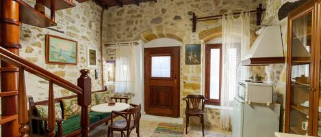 A rustic stone interior with a wooden staircase, a dining area with a table and chairs, a kitchen with a refrigerator, and a door with a window.