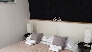 A neatly made bed with a beige quilt, two pillows, and a folded towel. There is a bedside table with a lamp, a wall-mounted shelf above the bed, and a framed picture on the wall. - Palace Stafileo (Trogir)