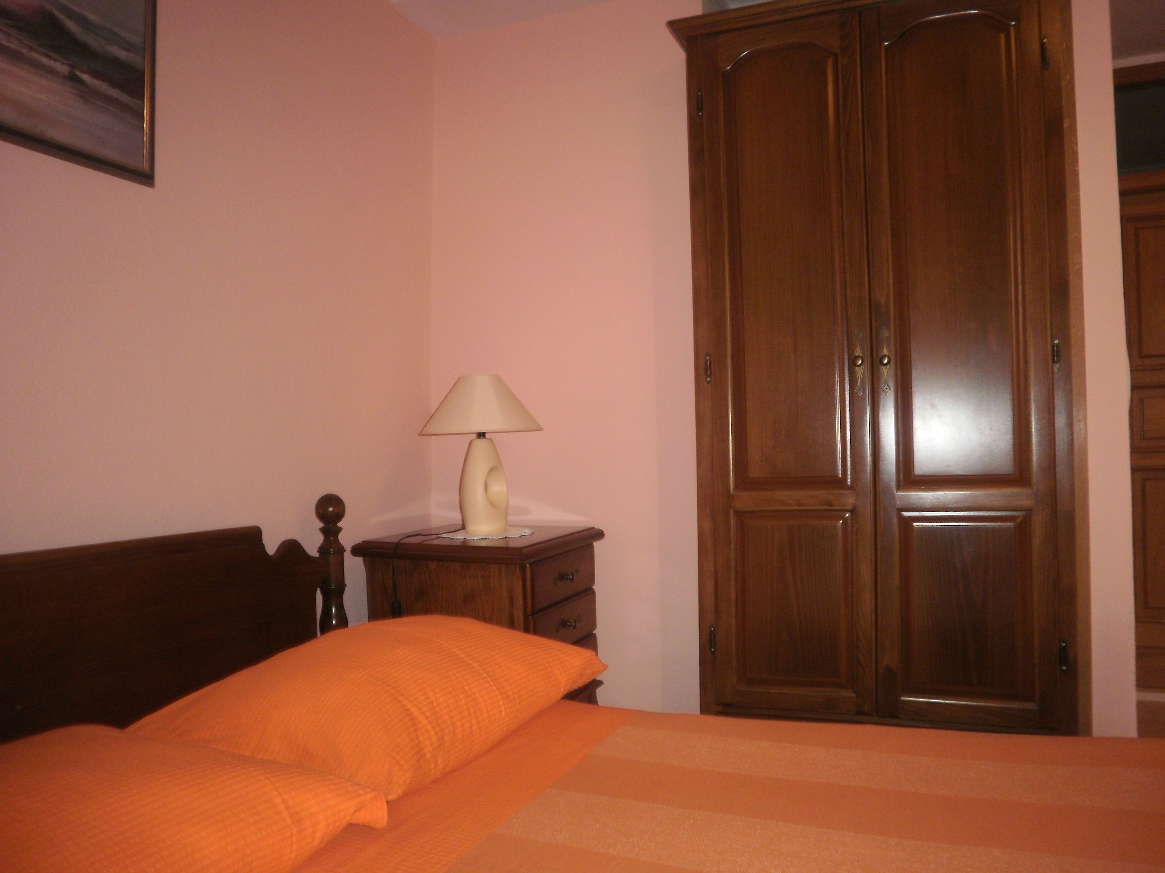 A bedroom with a bed, an orange blanket, a wooden wardrobe, and a bedside table with a lamp.